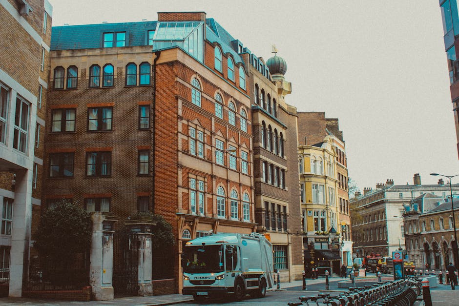 A street scene on Old Brompton Road featuring a mix of historic and modern buildings, including a prominent red-brick structure with arched windows and decorative façade detailing. In the foreground,