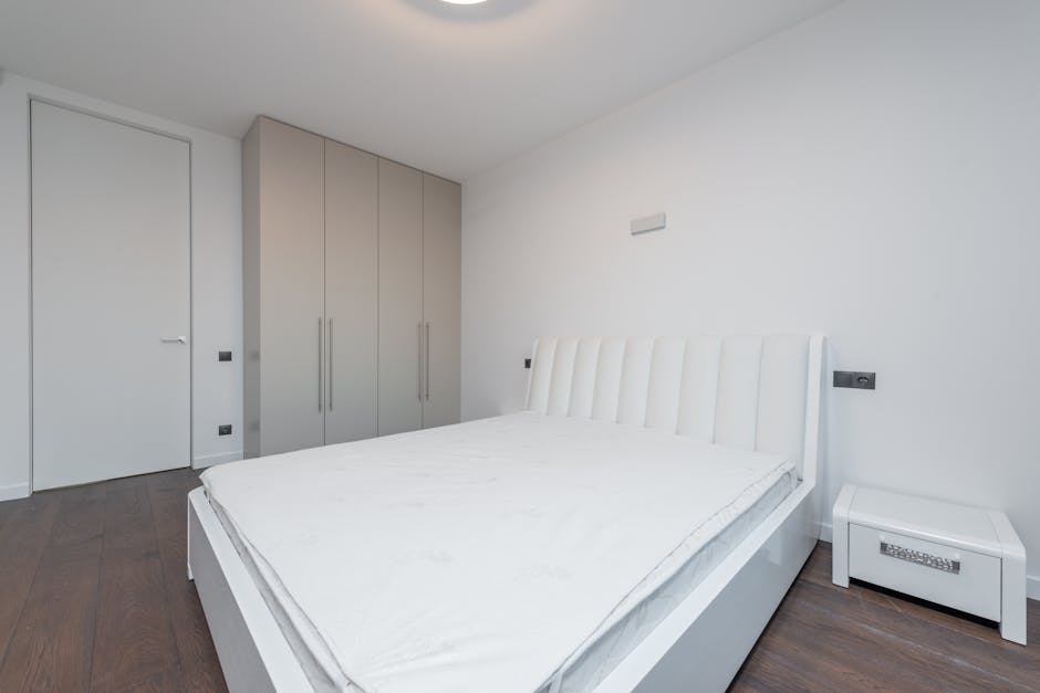 A bedroom featuring a large bed with a deep blue, fabric-upholstered, vertically tufted headboard, positioned against a soft pink accent wall. The mattress on the bed is viewed from above and appears